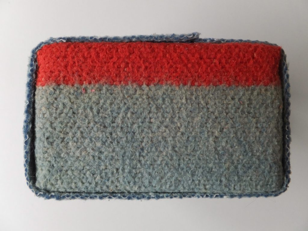  Enkel karakter van wollen deken uit het kunstwerk ‘Geborgen herinneringen’ door Jeanne de Bie met de kleuren grijs en rood tegen lichte achtergrond.